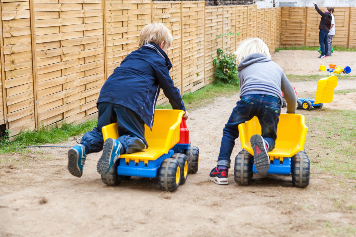 Zwei Kinder fahren im Freien auf großen gelben Spielzeug-LKWs nebeneinander her.