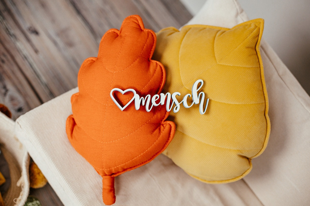 Zwei Kissen in Blattform, eines orange und eines gelb, mit einem Schriftzug ‚Herzmensch‘ aus Holz davor.