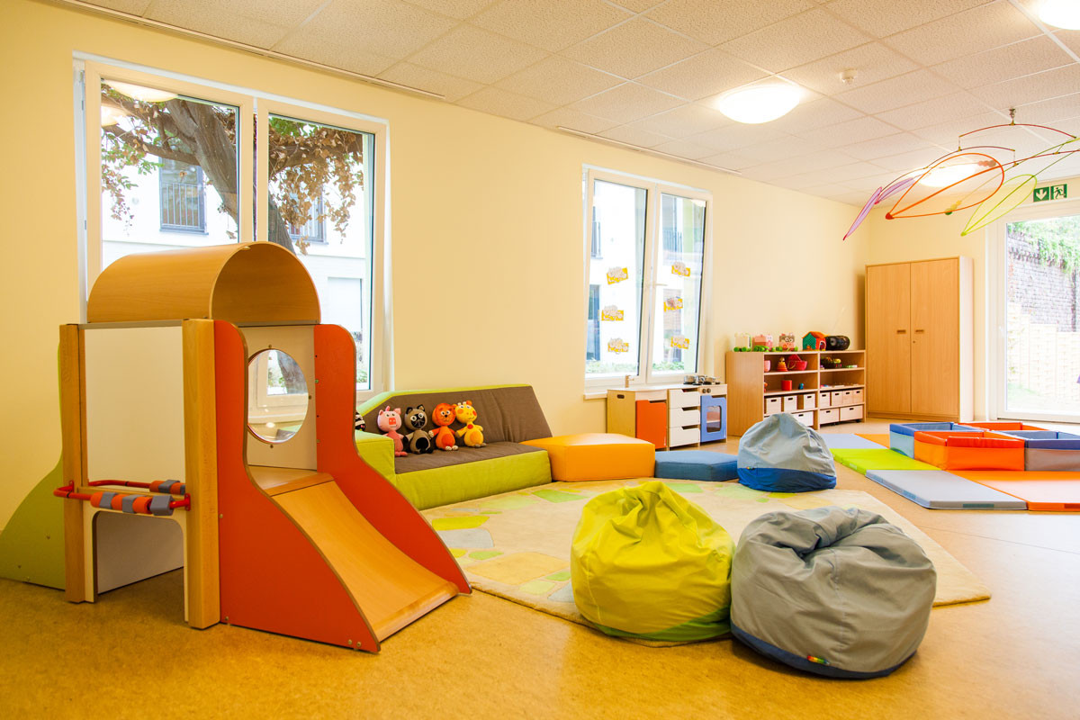 Heller Gruppenraum mit kleiner Rutsche, bunten Sitzsäcken, Spielmatten und Sofa mit Stofftieren.