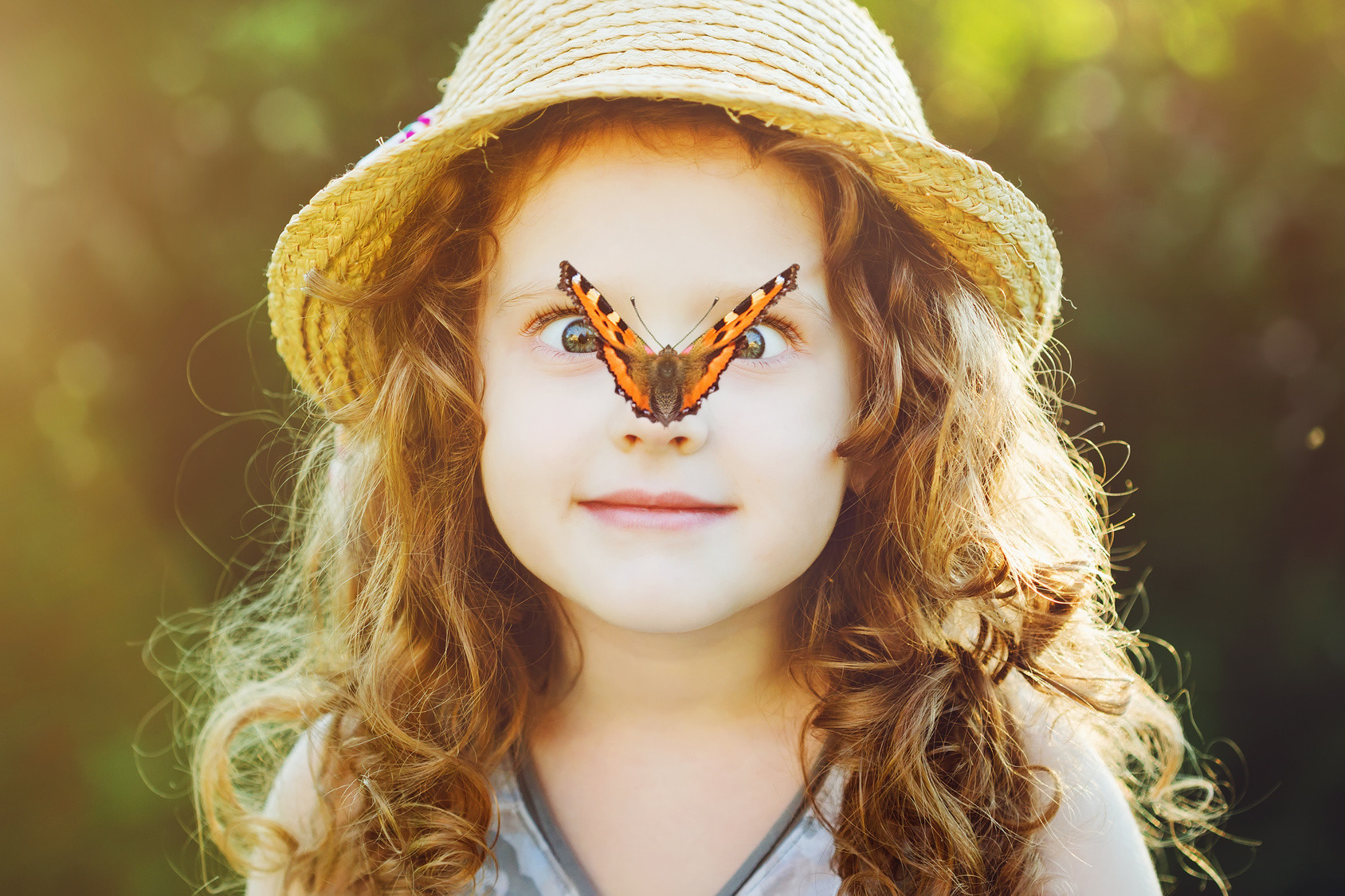 Ein kleines Mädchen mit lockigen Haaren und Strohhut, auf dessen Nase ein bunter Schmetterling sitzt.