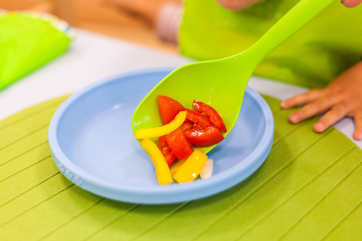 Mit einem grünen Löffel werden Tomaten- und Paprikastücke auf einen blauen Teller portioniert.