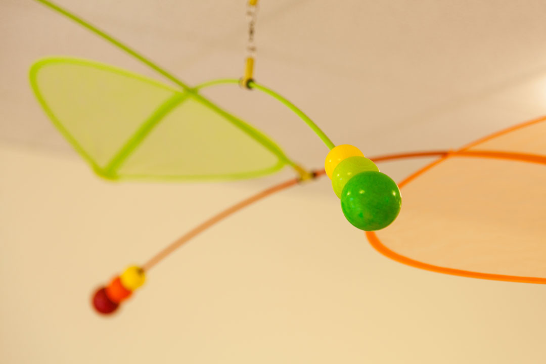 Buntes Mobile mit grünen und orangefarbenen Flügeln sowie fabrigen Kugeln hängt von der Decke.