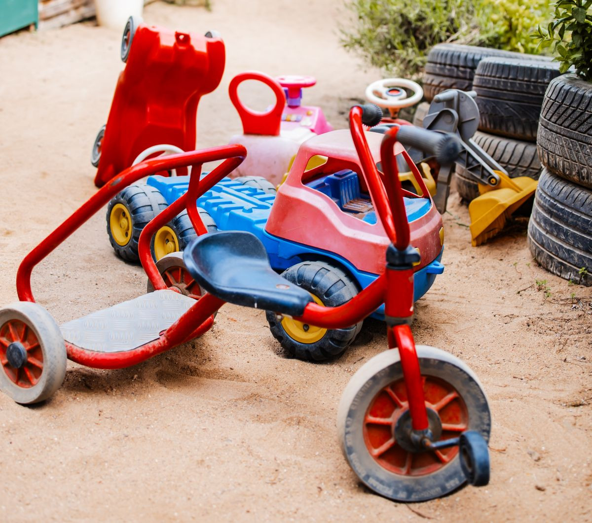Mehrere bunte Kinderfahrzeuge, darunter ein Dreirad und Spielautos, stehen auf sandigem Boden.