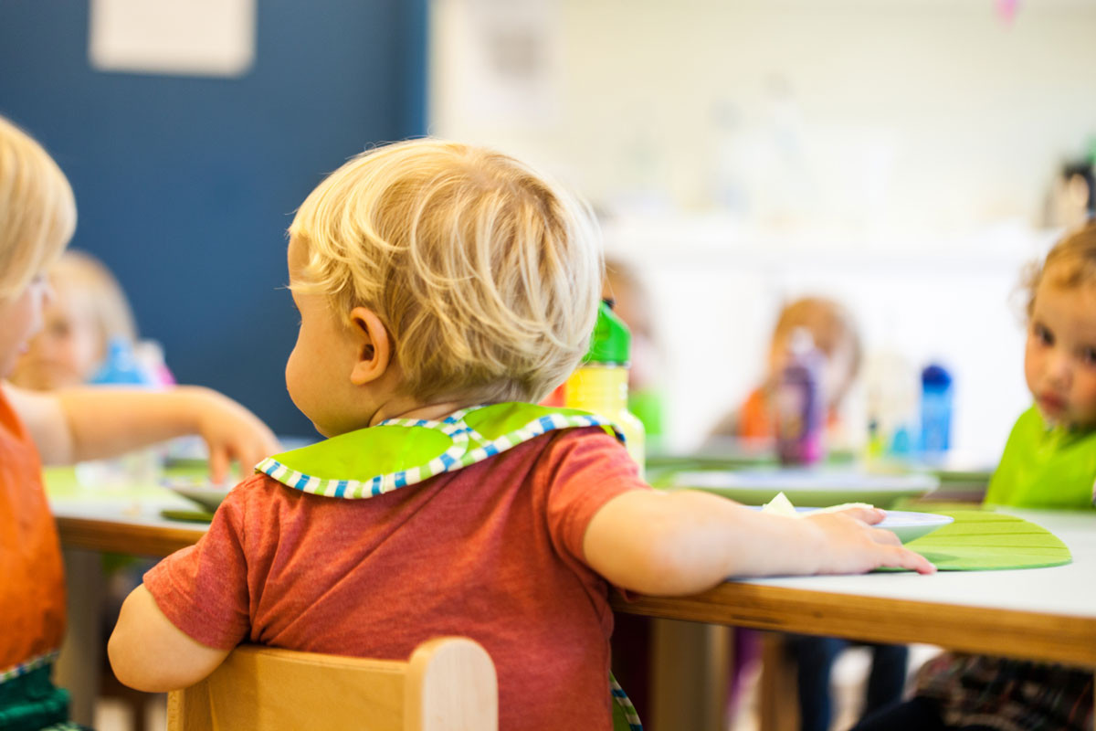 Kind mit Lätzchen sitzt am Tisch in der Kita, umgeben von weiteren Kindern.