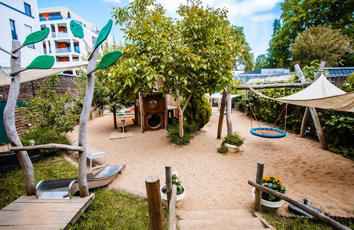 Spielplatz im Freien mit Sandboden, Bäumen und Holzspielgeräten: eine Rutsche, eine Kletter- und Röhrenanlage sowie eine runde Netzschaukel unter einem Sonnensegel.