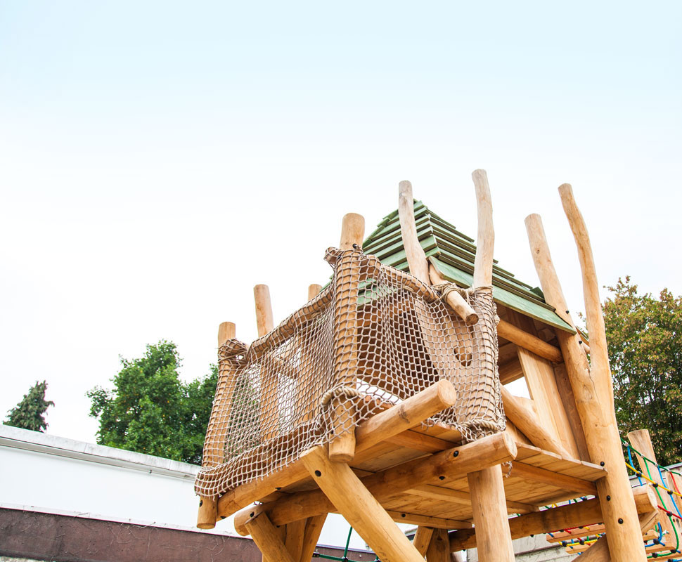 Holz-Kletterturm mit grünem Dach und Netzumrandung auf dem Spielplatz.