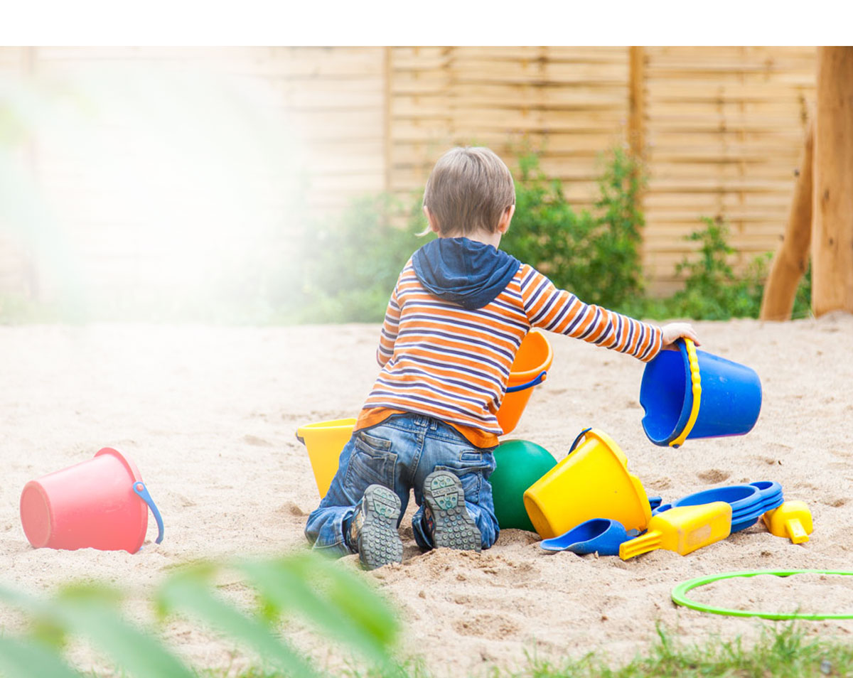 Ein Kind in Jeans und gestreiftem Pullover spielt im Sandkasten mit bunten Eimern und Schaufeln.
